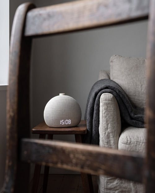 Halcyon wake-up light on a wooden stool beside an armchair, display reading 15:08